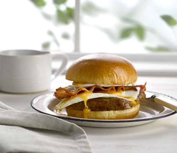 Breakfast Brioche burger with sausage bacon and egg, and a cup of coffee in the background